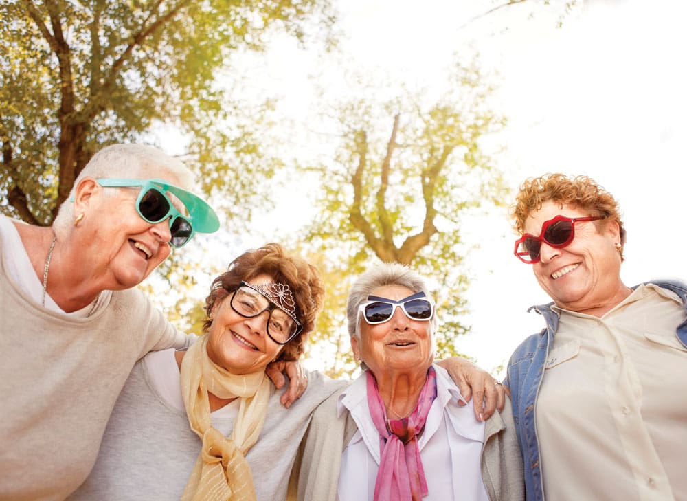 Group of older women put their arms around each other and pose outside in fun sunglasses 