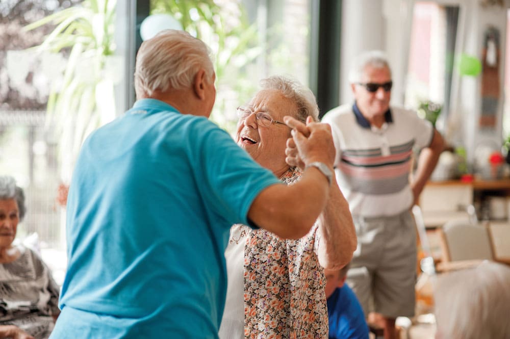 Senior living male and female share a dance surrounded by friends in the activity room 