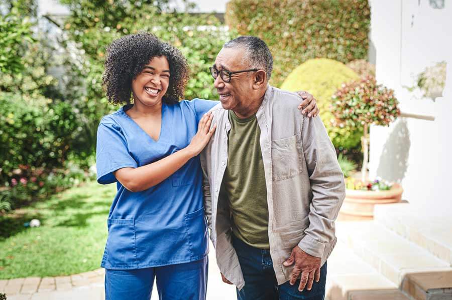A smiling caregiver warmly assisting and chatting with a happy senior man in a sunny outdoor setting.